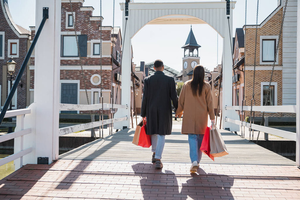 back view of interracial couple in coats holding hands and walking on bridge with shopping bags - Фото, зображення