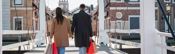 back view of trendy multiethnic couple with shopping bags holding hands on bridge, banner - Zdjęcie, obraz