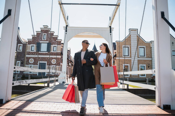 stylish interracial couple smiling at each other and walking with shopping bags across bridge - Фото, изображение