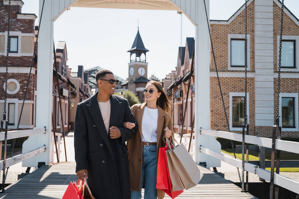 fashionable and happy multiethnic couple with shopping bags on city bridge - Fotó, kép