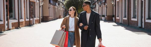 sonriente pareja multiétnica en abrigos de moda y gafas de sol que llevan bolsas de compras, pancarta - Foto, Imagen