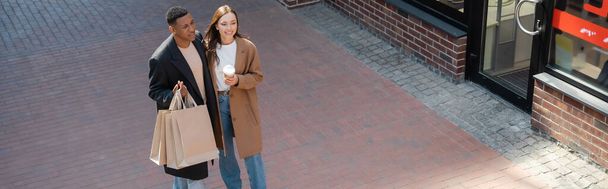 cheerful interracial couple in stylish coats holding takeaway drink and shopping bags on city street, banner - Foto, imagen
