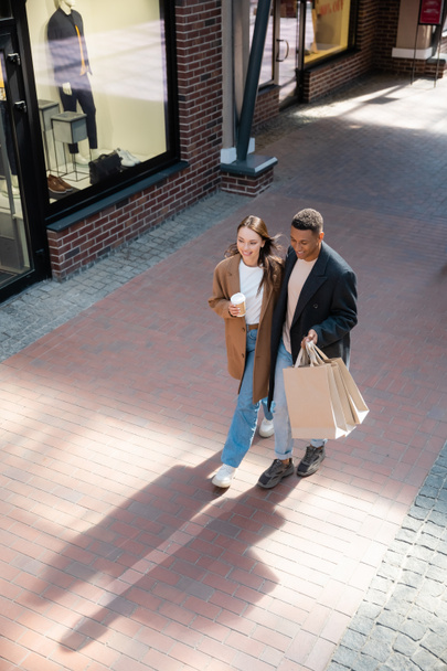 high angle view of happy and fashionable multiethnic couple with shopping bags and paper cup walking near shops on street - Foto, Imagem