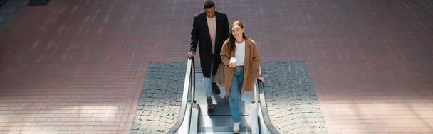 vue grand angle du couple multiethnique dans des manteaux à la mode tenant tasse en papier et sacs à provisions sur escalator, bannière - Photo, image