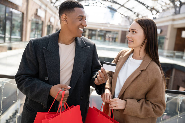 feliz africano americano hombre mostrando tarjeta de crédito a complacido joven novia sosteniendo bolsas de compras - Foto, imagen