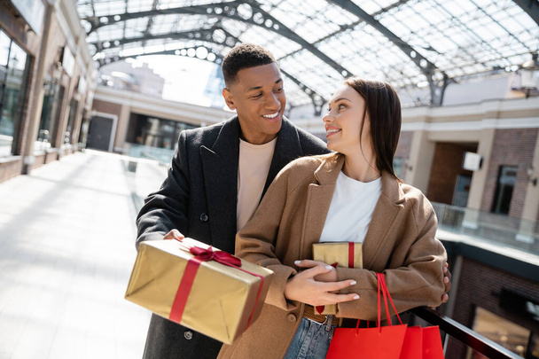 uomo afro-americano alla moda che dà il nuovo regalo di anno alla fidanzata felice con borse della spesa - Foto, immagini