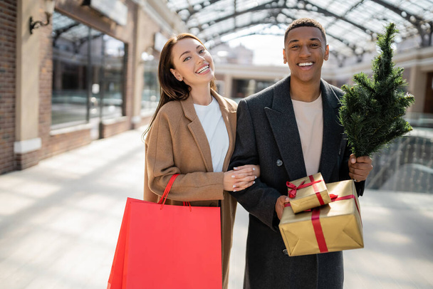 allegra coppia interrazziale con borse della spesa e scatole regalo con piccolo albero di Natale guardando la fotocamera vicino al centro commerciale - Foto, immagini
