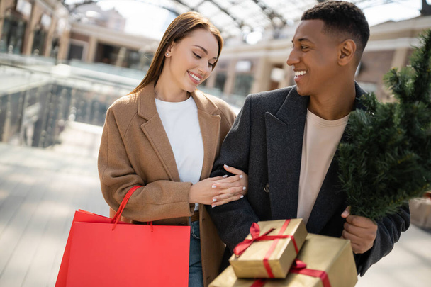 小さなクリスマスツリーとギフトボックスを持つスタイリッシュなアフリカ系アメリカ人男性見て楽しいですガールフレンドでショッピングバッグ - 写真・画像