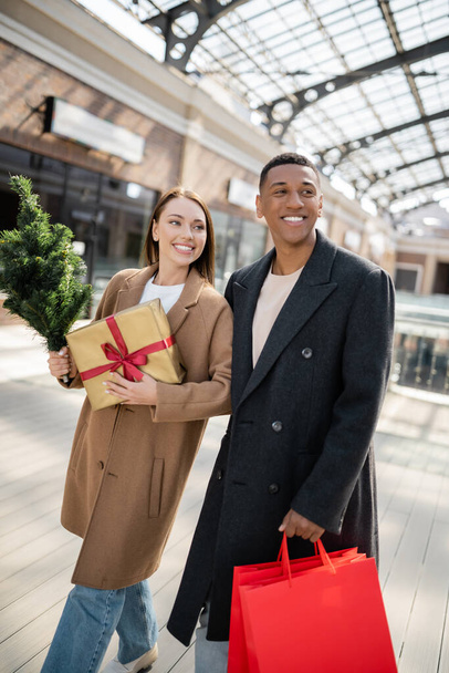 divatos nő ajándékdobozzal és kis karácsonyfa néz félre közel mosolygó afro-amerikai férfi bevásárló táskák - Fotó, kép