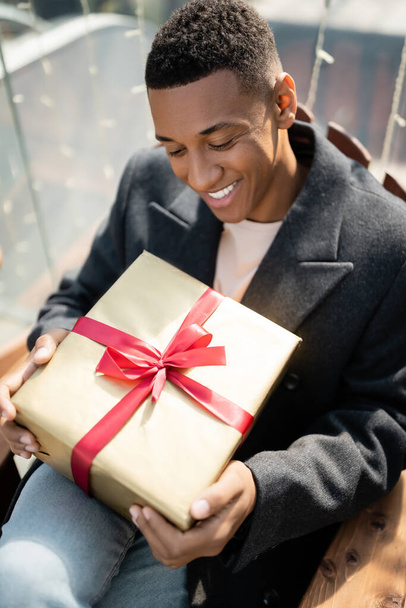 happy african american man in coat sitting with gift box outdoors - Photo, Image