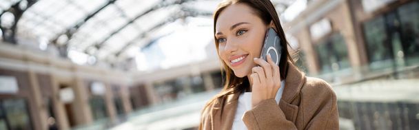 stylish brunette woman smiling during conversation on cellphone outdoors, banner - Foto, immagini