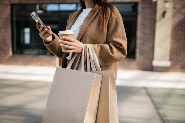 cropped view of woman using smartphone and holding shopping bags with paper cup on city street - Foto, Imagen