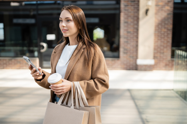 smiling woman in beige coat holding smartphone while standing with shopping bags and paper cup on blurred street - Fotografie, Obrázek