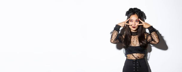Красивая азиатка в костюме для вечеринки на Хэллоуин смотрит в верхний левый угол, делает мирные жесты над глазами, стоя в готическом кружевном платье с черным венком на белом фоне. - Фото, изображение