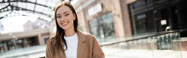 brunette woman in beige coat smiling at camera on blurred city street, banner - Foto, immagini