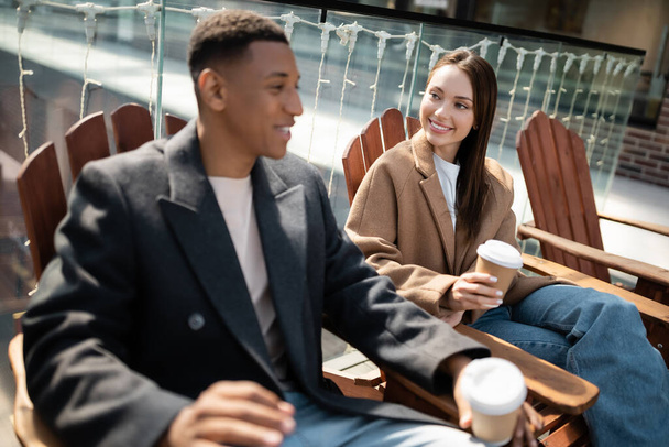 smiling interracial couple in coats sitting with paper cups on wooden seats outdoors - Photo, image