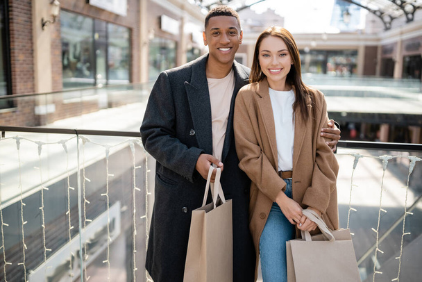 cheerful multiethnic couple in trendy coats holding shopping bags and looking at camera near blurred mall - Foto, imagen