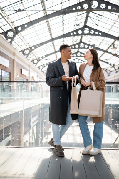 longitud completa de la pareja multiétnica de moda con bolsas de compras hablando cerca de centro comercial borrosa - Foto, imagen