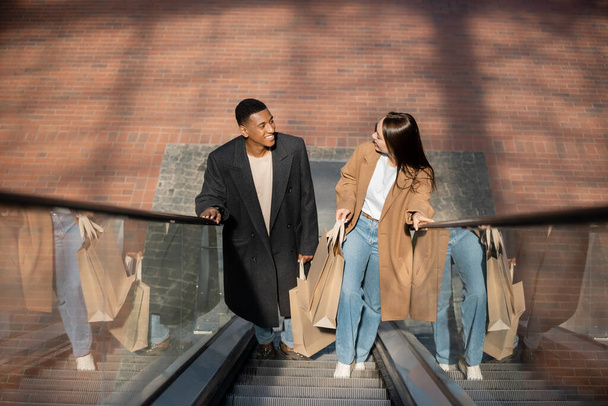high angle view of fashionable interracial couple with shopping bags smiling at each other on escalator - Foto, immagini