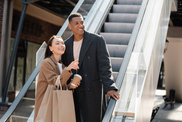 cheerful woman with paper cup and shopping bags pointing with finger near trendy african american man on escalator - Foto, Bild
