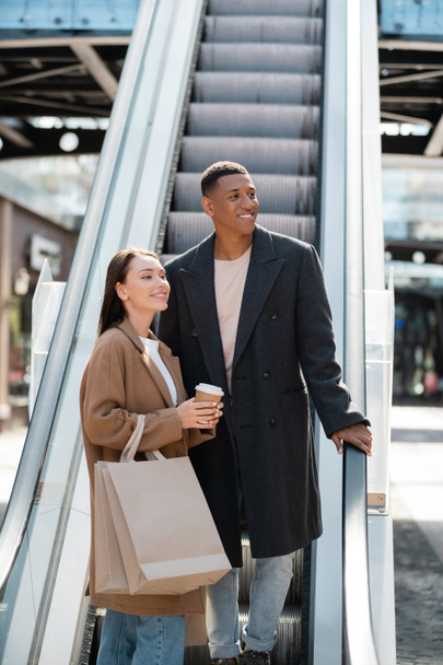 Afrikanisch-amerikanischer Mann in stylischem Mantel schaut bei junger Freundin mit Coffee to go und Einkaufstüten auf Rolltreppe weg - Foto, Bild