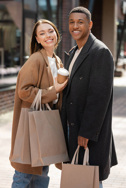 pleased young woman with takeaway drink and shopping bags looking at camera near trendy african american man - Foto, imagen