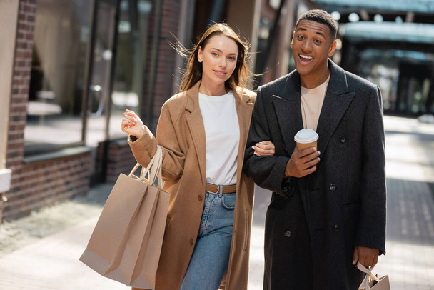 Komik ve neşeli Afrikalı Amerikalı adam alışveriş çantalı genç kız arkadaşının yanındaki kameraya bakıyor. - Fotoğraf, Görsel