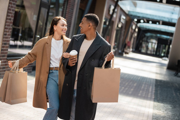 модная многонациональная пара в пальто, держащая сумки и улыбающаяся друг другу на городской улице - Фото, изображение