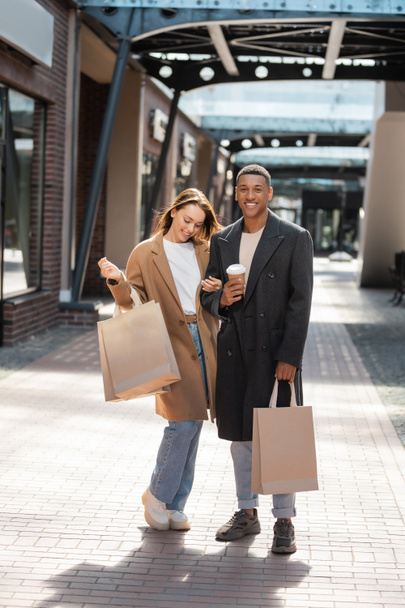 hombre afroamericano con estilo con bolsas de compras y taza de papel mirando a la cámara cerca de novia alegre - Foto, imagen