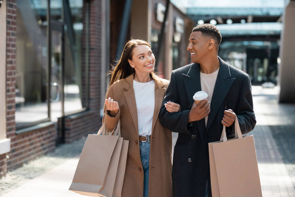 pleased multiethnic couple in fashionable clothes looking at each other while carrying shopping bags on street - Foto, imagen