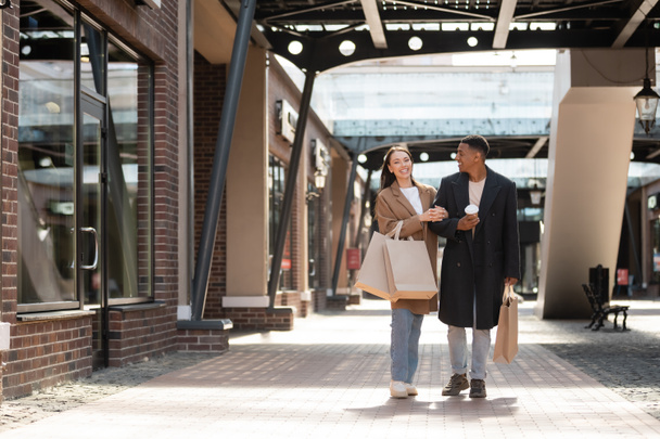 happy interracial couple in trendy coats walking with purchases along shops on urban street - Foto, Imagen