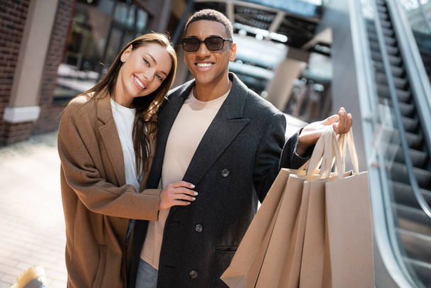 happy and stylish woman hugging african american boyfriend in sunglasses holding shopping bags on street - Фото, зображення