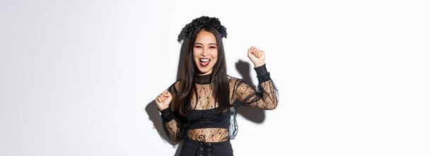 Cadılar bayramı partisinde eğlenen genç Asyalı bir kadının resmi. Neşeli ya da muzaffer olarak elini kaldırıyor. Cadı gotik elbisesi giyiyor, beyaz arka planda duruyor.. - Fotoğraf, Görsel