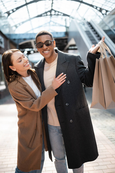 vrolijk jong vrouw kijken naar winkelen tassen in de hand van trendy Afrikaans amerikaanse man in een zonnebril  - Foto, afbeelding