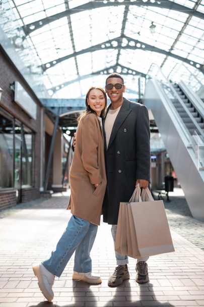 longitud completa de hombre afroamericano sonriente en gafas de sol sosteniendo bolsas de compras y abrazando novia de moda - Foto, Imagen
