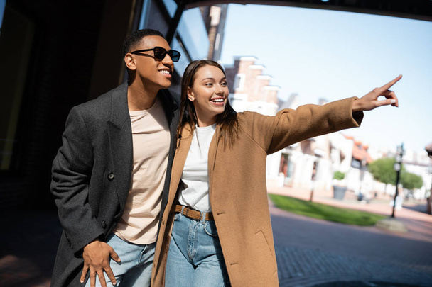 excited woman in coat looking away and pointing with finger near african american man in sunglasses - Fotoğraf, Görsel