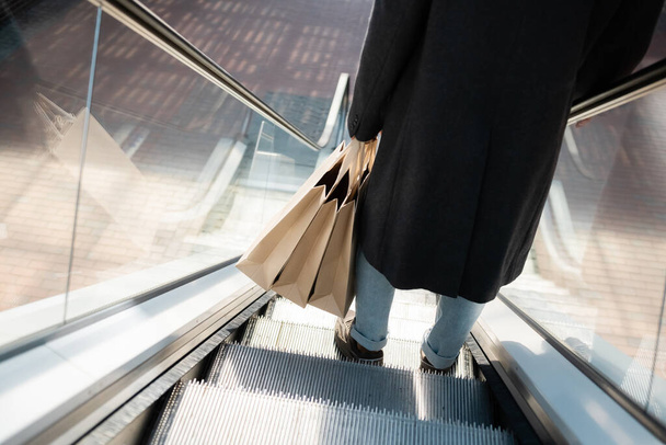 άποψη υψηλής γωνίας του περικοπή άνθρωπος σε παλτό κρατώντας τσάντες ψώνια στις κυλιόμενες σκάλες - Φωτογραφία, εικόνα