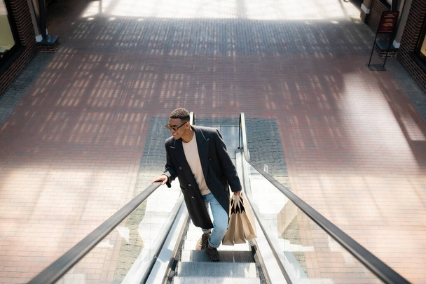high angle view of smiling and trendy african american man with shopping bags on escalator - Fotoğraf, Görsel