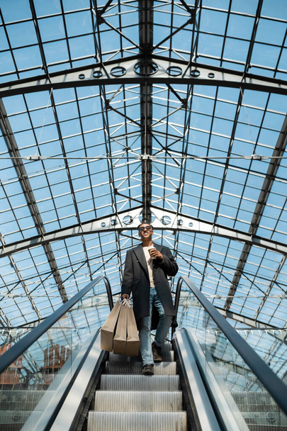 низький кут зору стильний афроамериканський чоловік з сумками для покупок і паперовою чашкою на ескалаторі під скляним дахом
 - Фото, зображення