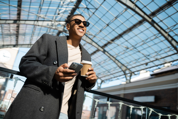 low angle view of trendy african american man with smartphone and coffee to go smiling under glass roof outdoors - Photo, Image
