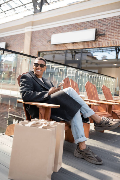 afro-amerikai férfi kabátban és napszemüvegben ül okostelefonnal közel bevásárlótáskák és bevásárlóközpont - Fotó, kép