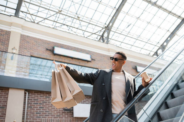podekscytowany Afroamerykanin mężczyzna w modnym płaszczu i okularach przeciwsłonecznych trzymając kawę iść i torby na zakupy na schodach ruchomych - Zdjęcie, obraz