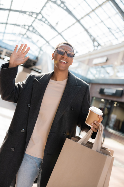 alegre afroamericano hombre con taza de papel y bolsas de compras agitando la mano en la calle de la ciudad - Foto, imagen