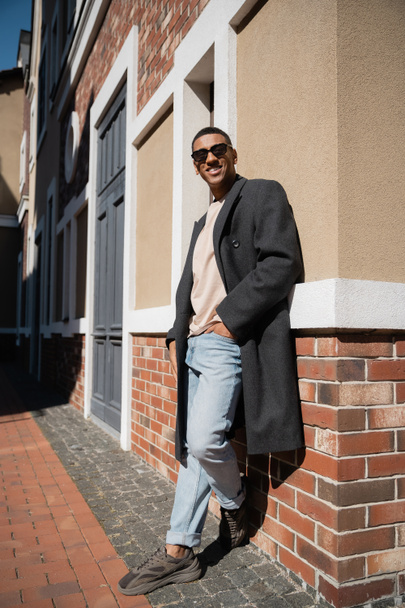 full length of african american man in coat and sunglasses standing with hand in pocket of jeans near building - Foto, Imagen