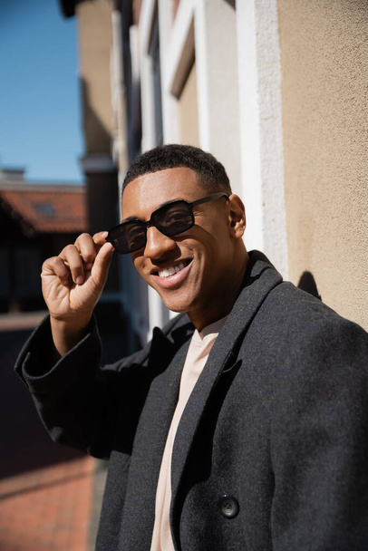 trendy african american man adjusting sunglasses and smiling at camera on city street - Photo, Image