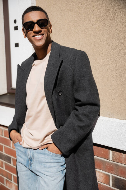 módní a šťastný africký Američan ve slunečních brýlích a kabátě stojí s rukama v kapsách džín v blízkosti budovy - Fotografie, Obrázek