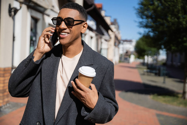 happy african american man in sunglasses and coat talking on smartphone and holding paper cup on street - 写真・画像