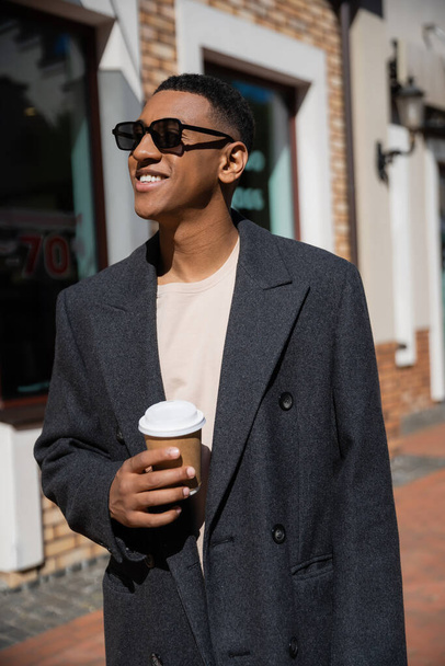 joyful and trendy african american man in sunglasses standing with paper cup and looking away - Фото, изображение