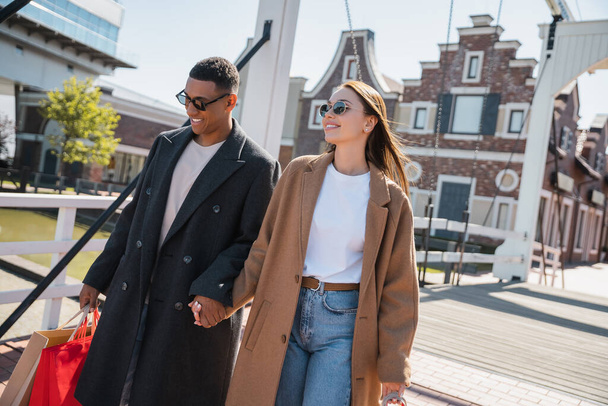 happy and fashionable couple in sunglasses holding hands while walking with shopping bags across city bridge - 写真・画像