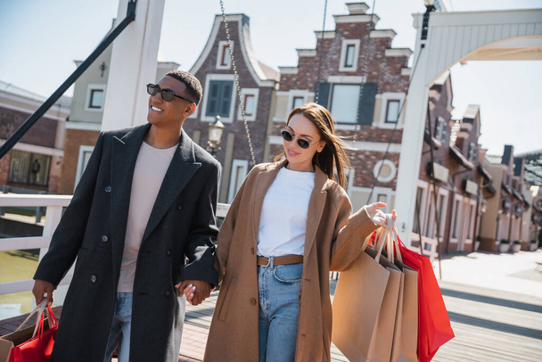 pleased and stylish interracial couple with shopping bags holding hands on city bridge - Foto, imagen